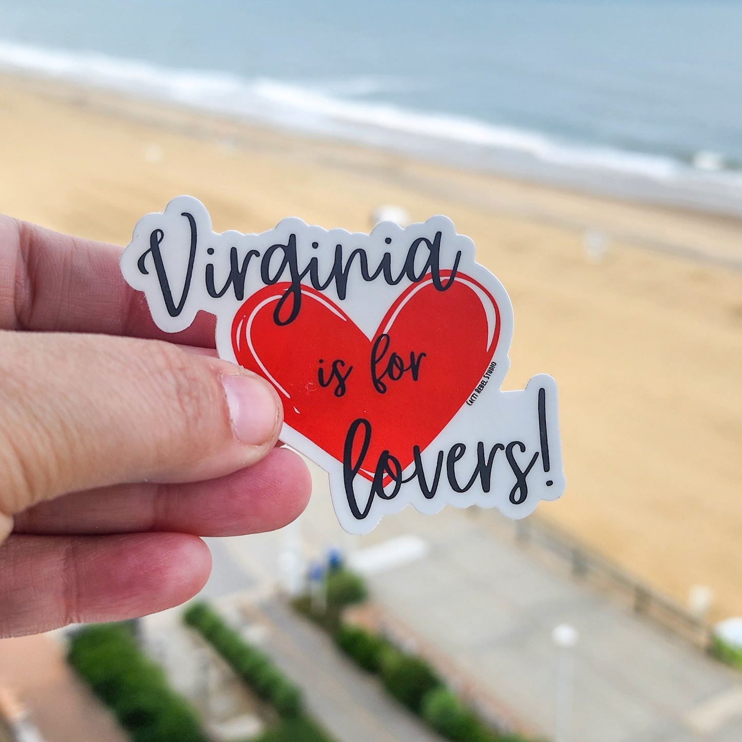 Hand holding a 'Virginia is for lovers!' sticker with a beach in the background