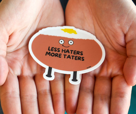 Sticker with a potato character and text 'LESS HATERS MORE TATERS' held between two hands.