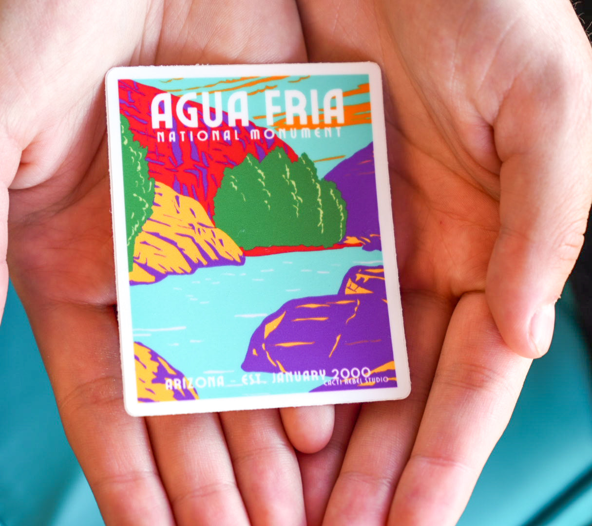 Colorful card with 'Agua Fria National Monument' design held between hands against a teal background