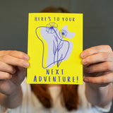 A person holding a yellow greeting card with a hand-drawn illustration of a cactus and flowers, and the text 'HERE'S TO YOUR NEXT ADVENTURE!'.