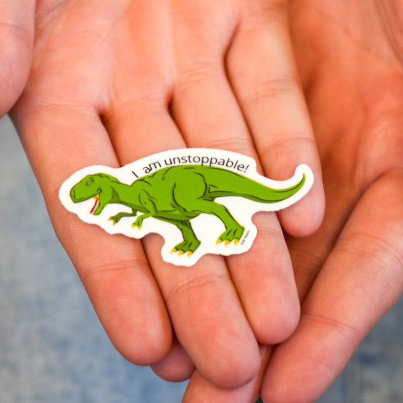 A hand holding a vinyl sticker of a t-rex dinosaur with the phrase 'I am unstoppable!' printed in vibrant colors.