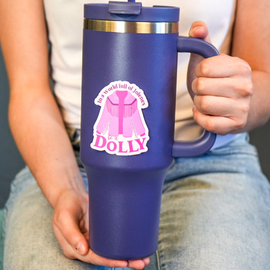 A vinyl die cut sticker with the text 'In a world full of Jolene's be a Dolly' held by a person, predominantly purple in color with a pink graphic and white text.