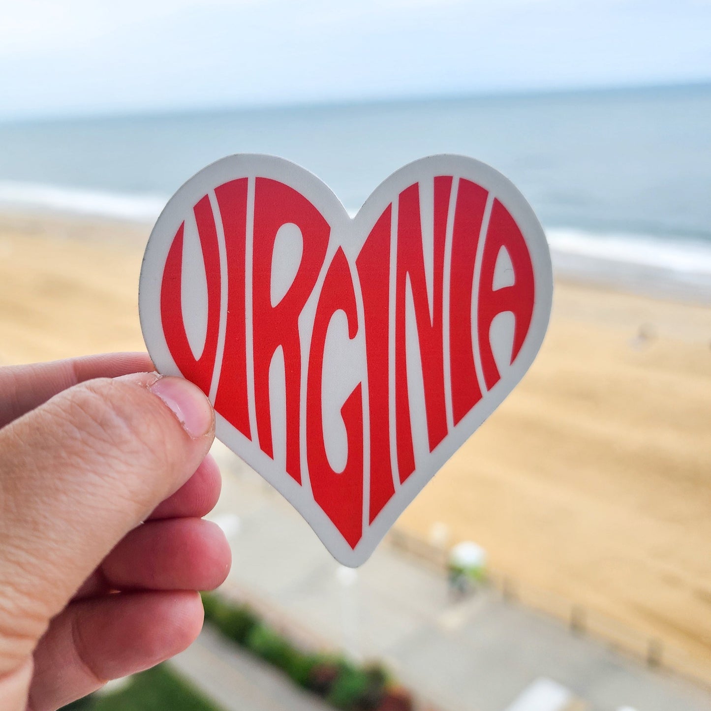 Heart-shaped 'Virginia' sticker held by a hand with a blurred beach background