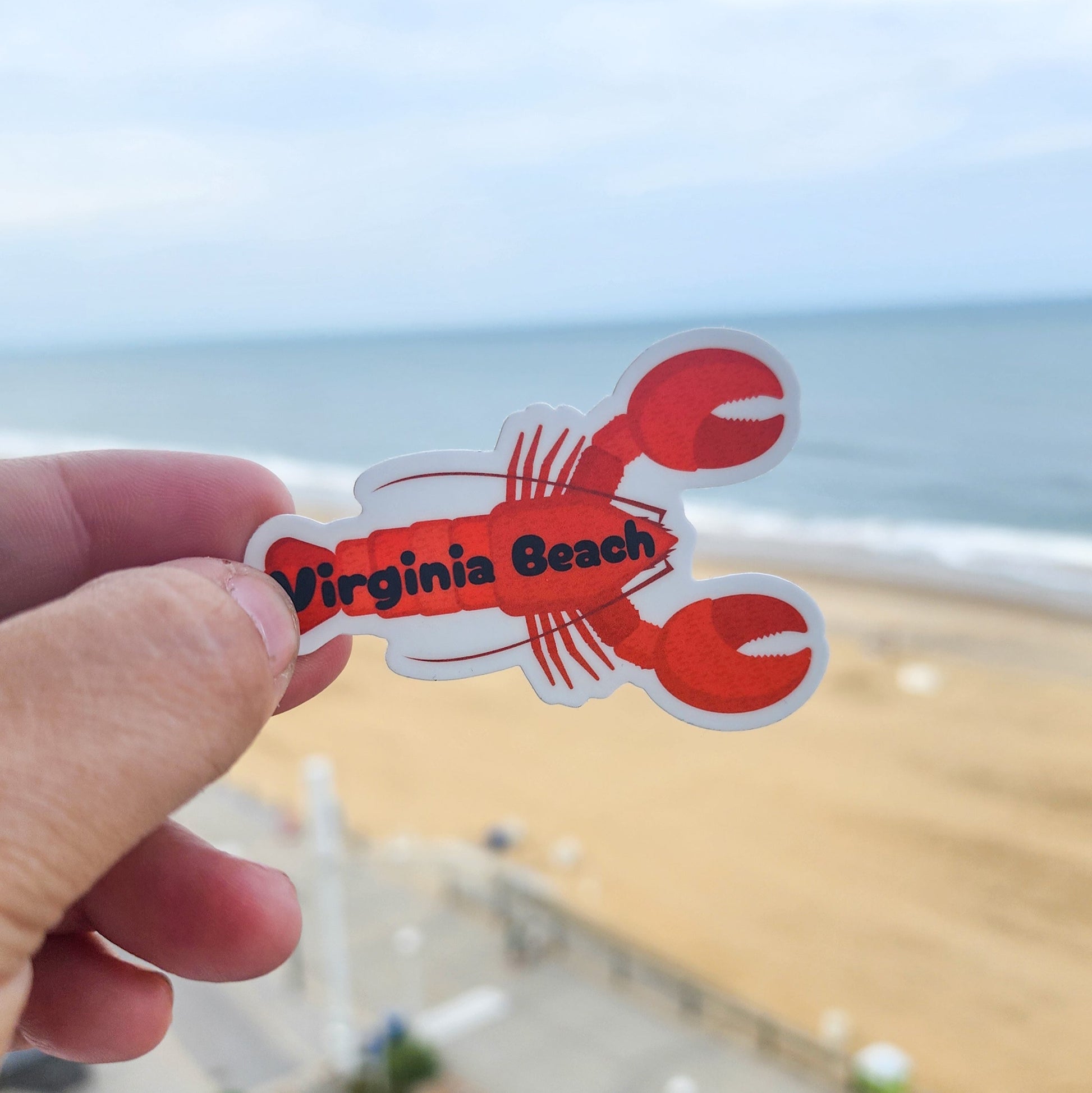 Red 'Virginia Beach' lobster sticker held up with a beach in the background