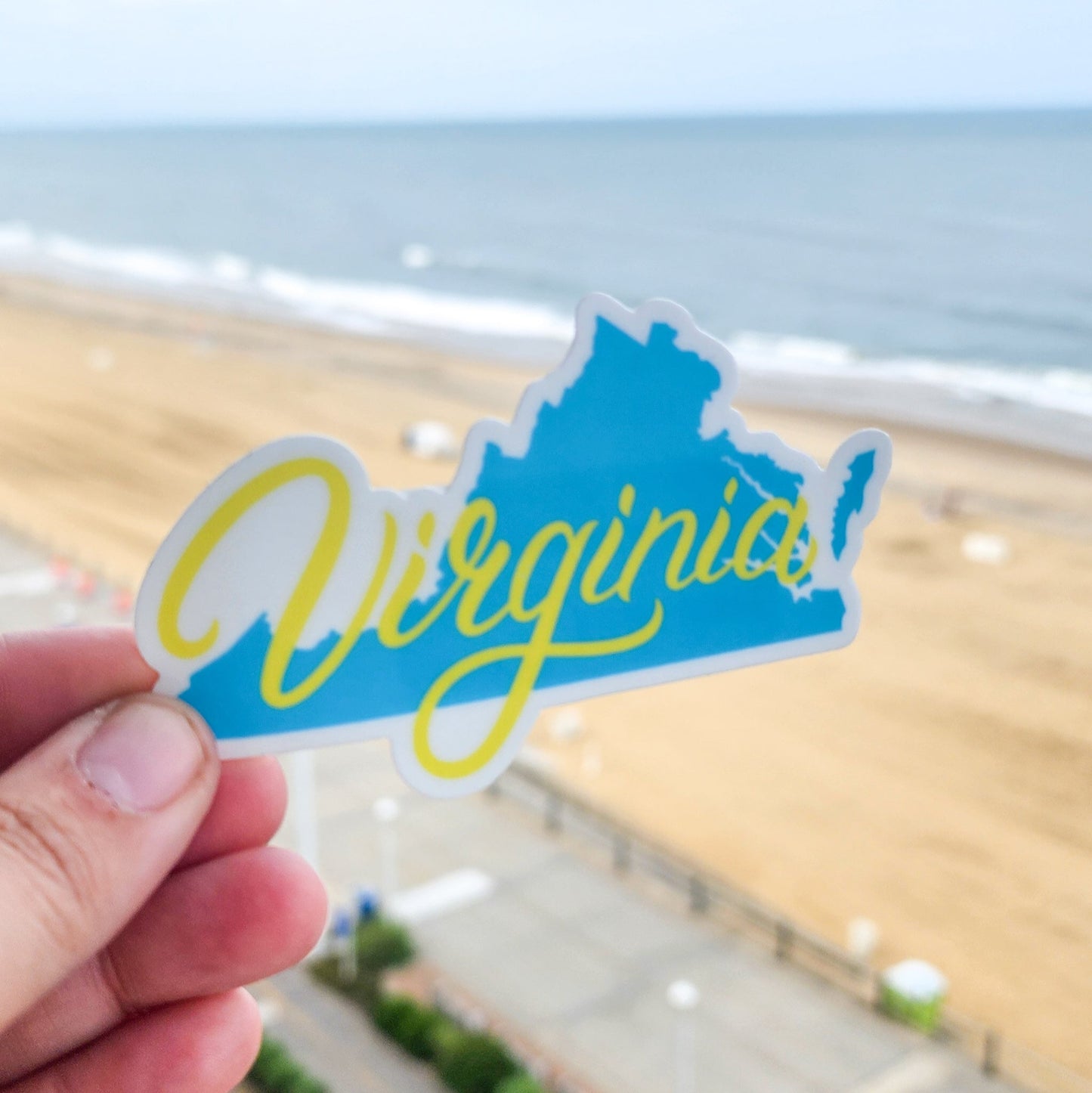 Hand holding a blue and yellow 'Virginia' sticker with a beach and ocean view in the background.