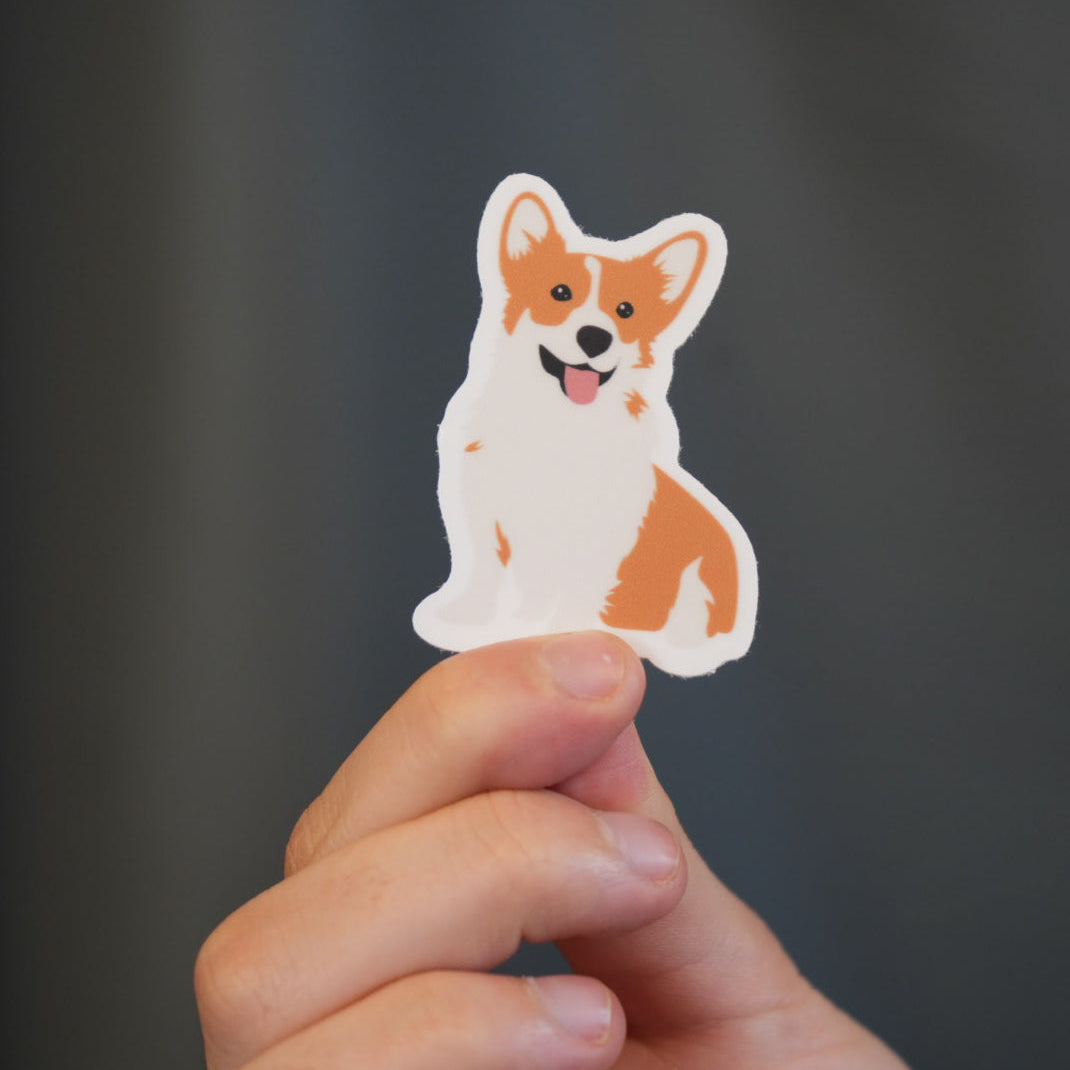A vinyl die cut sticker of a corgi dog held between fingers, featuring vibrant colors and a hand-drawn illustration.
