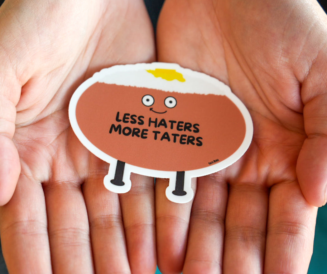 Sticker with a potato character and text 'LESS HATERS MORE TATERS' held between two hands.