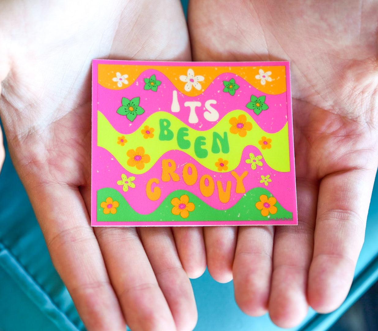 Colorful square sticker with 'It's Been Groovy' text held between two hands against a blue background