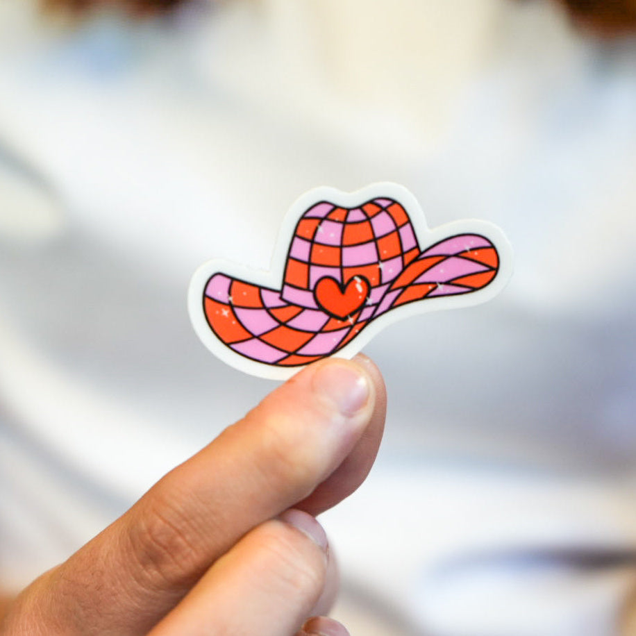 A vinyl die cut sticker of a cowgirl hat with a pattern of disco hearts, held between fingers.