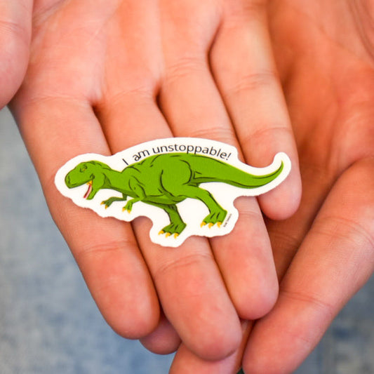 A hand holding a vinyl sticker of a t-rex dinosaur with the phrase 'I am unstoppable!' printed in vibrant colors.