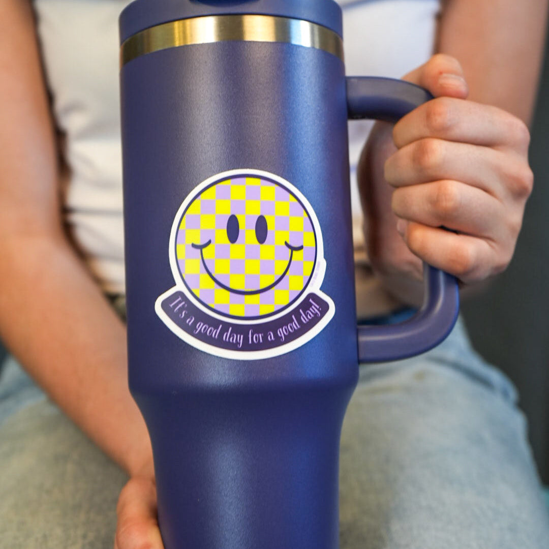 A person holding a blue water bottle with a vinyl die-cut sticker featuring a smiley face and the phrase 'It's a good day for a good day' in vibrant colors.