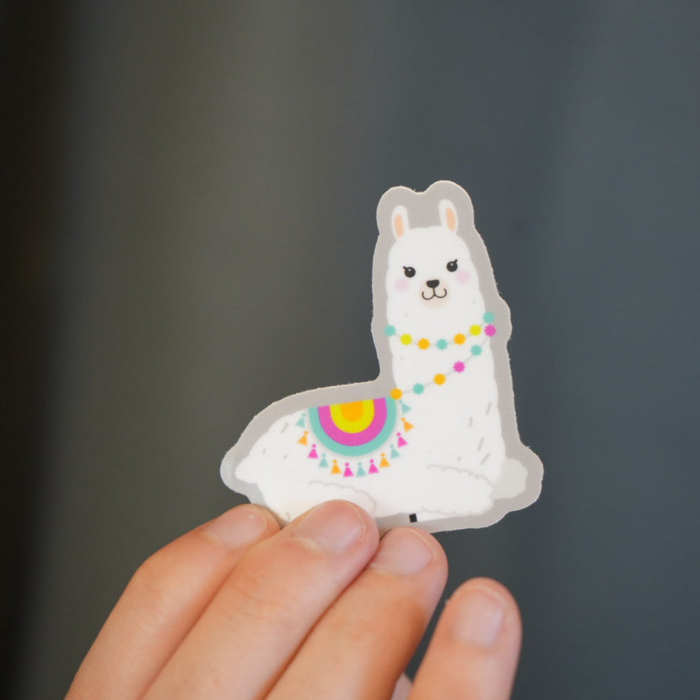 A vinyl die cut sticker of a white llama with colorful details, held between fingers.