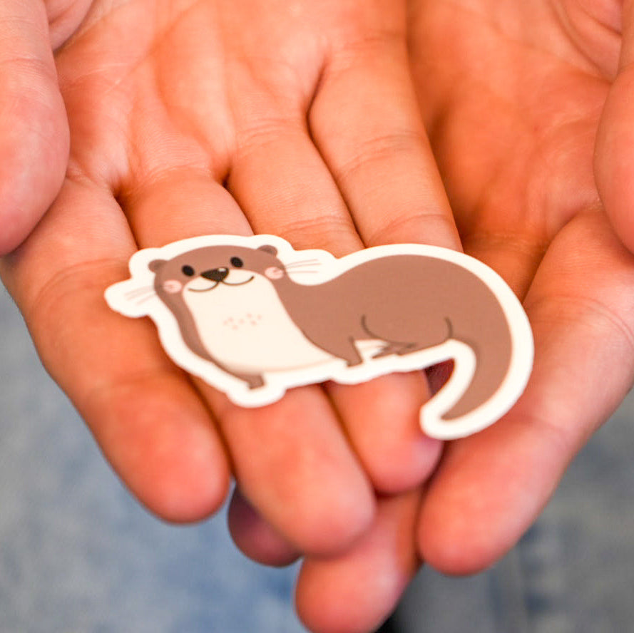 A vinyl die cut sticker of an otter, held in a person's palms.