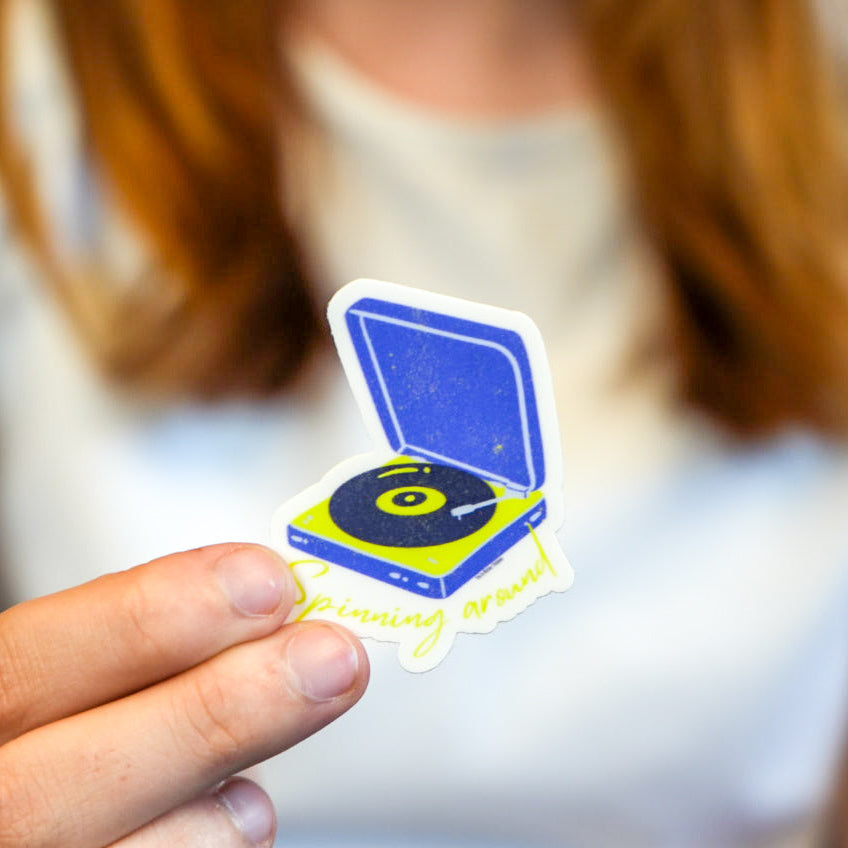 A hand holding a die-cut vinyl sticker of a blue record player with the text 'Spinning around' on it.