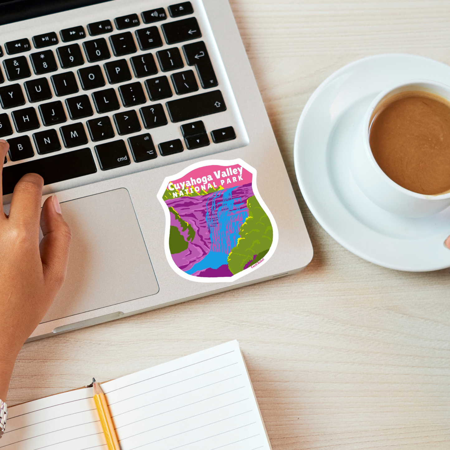 Person using a laptop with a coffee cup and tablet on a desk with a Cuyahoga Valley National Park sticker on the laptop