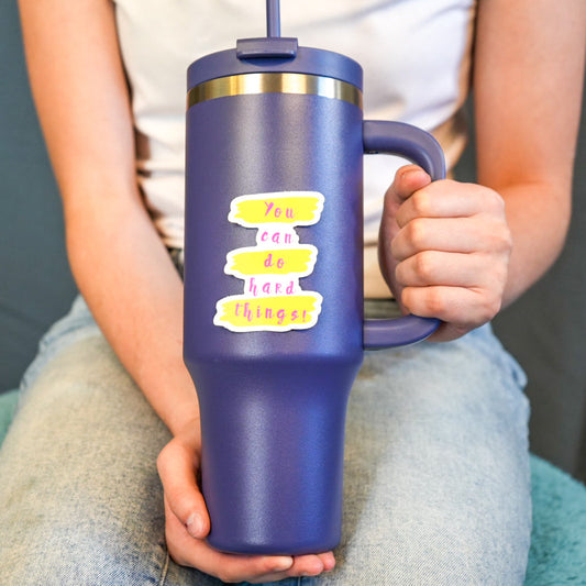 A person holding a navy-colored water bottle with a sticker that says 'You can do hard things' in yellow letters.