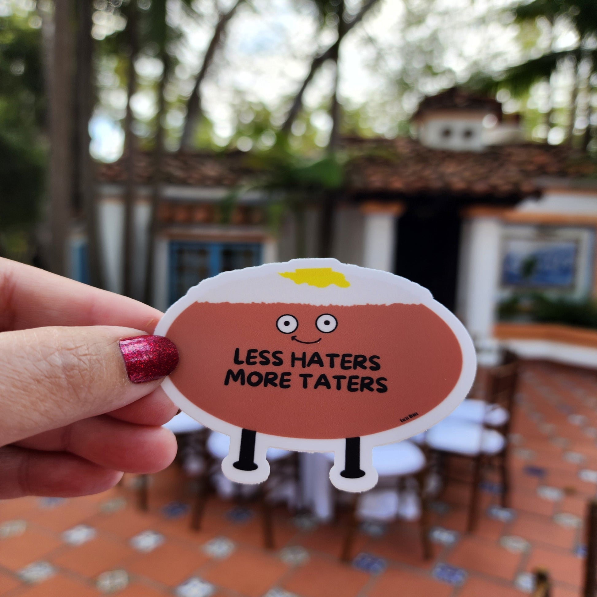 A die cut vinyl sticker with a cartoon illustration of a smiling potato with the phrase 'Less Haters More Taters' held in a hand, with a background of a patio and furniture.
