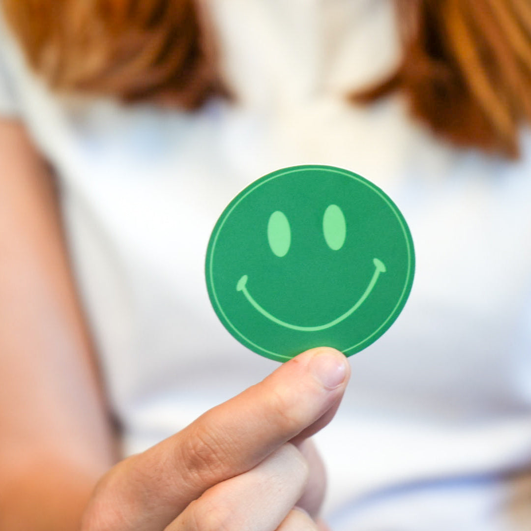 A person holding a round green sticker with a smiley face drawing in a indoor setting.