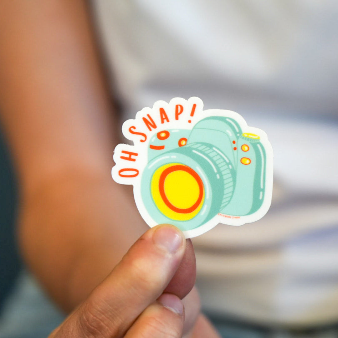 A vinyl die cut sticker held between fingers, featuring a colorful design of a camera with the phrase 'OH SNAP!' across the top.